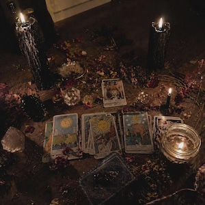 May include: A dimly lit scene featuring tarot cards, lit candles, crystals, and scattered rose petals. The cards are arranged on a wooden surface, with several lit candles casting shadows. The overall aesthetic suggests a mystical or spiritual theme.