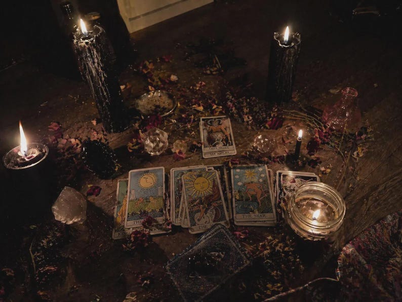 May include: A dimly lit scene featuring tarot cards, lit candles, crystals, and scattered rose petals. The cards are arranged on a wooden surface, with several lit candles casting shadows. The overall aesthetic suggests a mystical or spiritual theme.