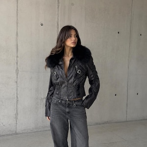 Könnte beinhalten: Eine schwarze Jacke mit Kunstfellkragen, Frontreißverschluss und Schnallendetails auf der Brust, kombiniert mit einer weit geschnittenen anthrazitfarbenen Jeans. Die Jacke hat lange Ärmel mit Schnürdetails an den Seiten und eine taillierte Passform.