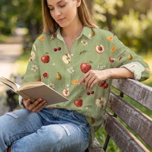 Puede incluir: Una camisa de manga larga abotonada de color verde claro con un estampado integral de varias frutas, incluyendo manzanas rojas, manzanas en rodajas, cerezas, fresas, rodajas de naranja, higos y peras, intercaladas con pequeñas flores blancas y hojas verdes.