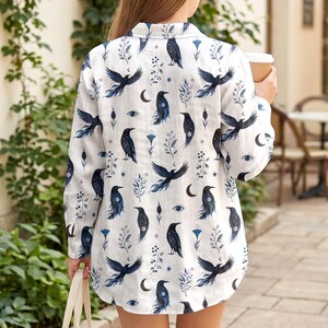 May include: White button-down shirt with a repeating pattern of black crows, crescent moons, and blue floral and eye designs. The long-sleeved shirt is made of a lightweight fabric.
