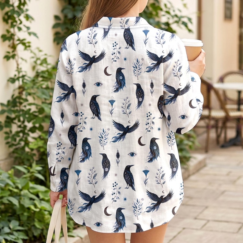 May include: White button-down shirt with a repeating pattern of black crows, crescent moons, and blue floral and eye designs. The long-sleeved shirt is made of a lightweight fabric.