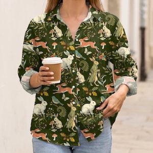 May include: A long-sleeved button-down shirt in a dark forest green color featuring an all-over print of white and brown rabbits, small mushrooms, green ferns, and colorful butterflies. The shirt has a classic collar and white buttons.