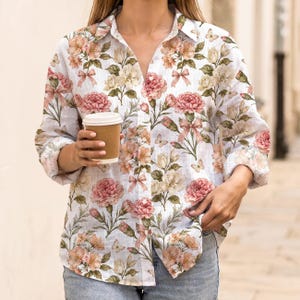 May include: A white floral shirt with a pattern of pink, peach, and cream flowers, bows, and butterflies. The shirt is worn with blue jeans. A brown paper coffee cup is held in the hand.