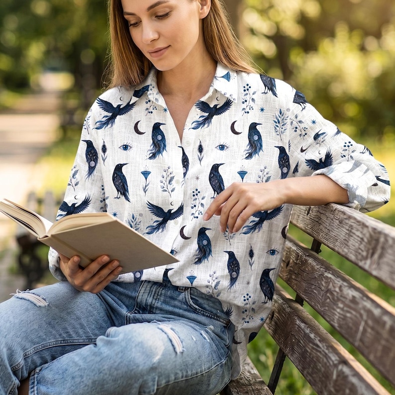 May include: White button-down shirt with a dark blue raven and celestial pattern. The long-sleeved shirt features a repeating design of ravens, crescent moons, and floral elements. The shirt is made of a lightweight fabric.