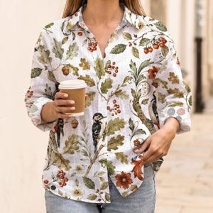Könnte beinhalten: Weißes Hemd mit botanischem Druck mit Eichenblättern, Eicheln, Beeren und Spechten. Das langärmlige Hemd hat einen Kragen. Eine Person hält eine braune Pappkaffeetasse.