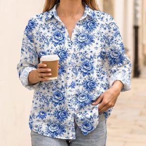 May include: A white shirt with a blue floral pattern. The shirt has a classic collar and long sleeves. The floral design includes roses and other flowers. A brown paper coffee cup is held in the hand.