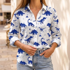 May include: A white long-sleeve button-down shirt featuring an all-over print of blue dinosaurs, including stegosauruses, brachiosauruses, and triceratops, along with small volcano icons and the text GRR! printed in a dark blue font.