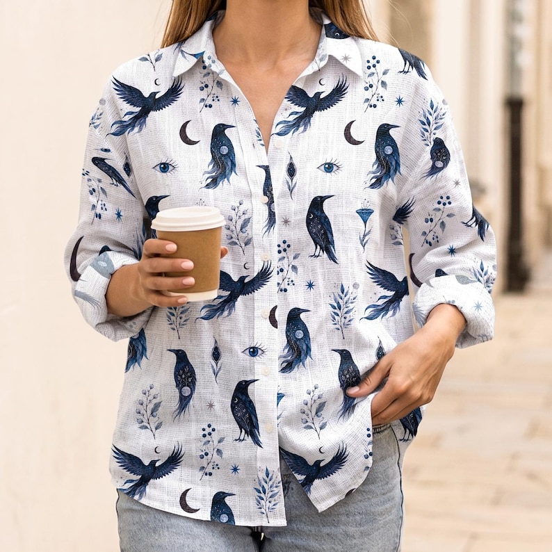 May include: White button-down shirt with a repeating pattern of dark blue ravens, crescent moons, and botanical elements. The shirt is long-sleeved and has a collared neckline. A person is holding a brown paper coffee cup.