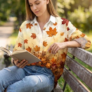 May include: A long-sleeved button-down shirt featuring a cream-colored base with an all-over print of autumn maple leaves in shades of orange, red, and brown. The shirt has a classic collar and is paired with light blue distressed denim jeans.