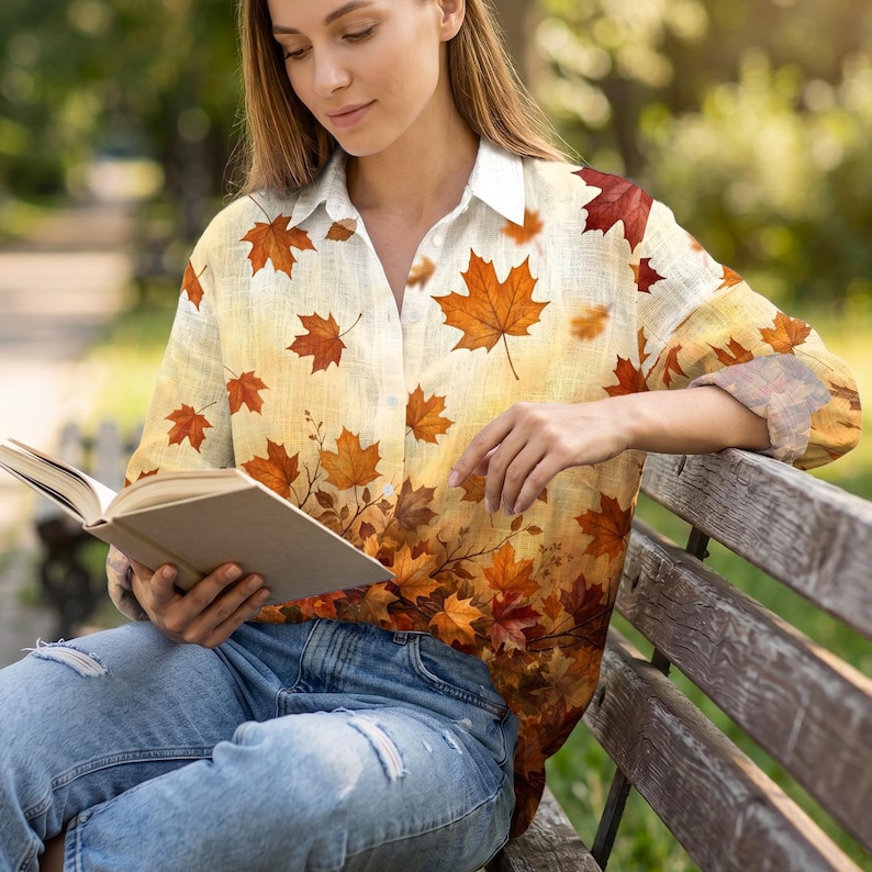 May include: A long-sleeved button-down shirt featuring a cream-colored base with an all-over print of autumn maple leaves in shades of orange, red, and brown. The shirt has a classic collar and is paired with light blue distressed denim jeans.