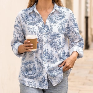 Puede incluir: Una camisa de manga larga con botones frontales que presenta un estampado toile de jouy azul sobre fondo blanco, con figuras históricas, árboles y motivos florales. La prenda cuenta con un cuello clásico y puños en las mangas.