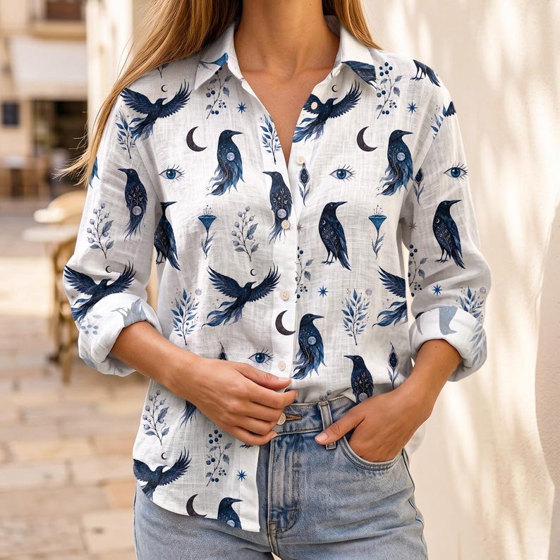 May include: White button-down shirt with a repeating pattern of dark blue crows, crescent moons, and floral elements. The long-sleeved shirt is paired with blue jeans. The shirt's design features detailed illustrations.