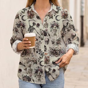 May include: A long-sleeved button-down shirt in a light beige color featuring an all-over print of skeletons, skulls, crescent moons, dark flowers, and mushrooms. The shirt has a classic collar, a front button placket, and cuffed sleeves.