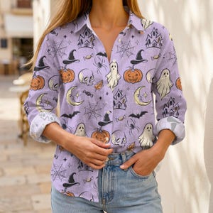 May include: A light purple long-sleeve button-down shirt featuring a repeating Halloween-themed pattern. The print includes illustrations of orange pumpkins, white ghosts, black witch hats, skulls, crescent moons, spider webs, and haunted houses.