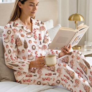 May include: A white long-sleeve button-down pajama set featuring a repeating pattern of nutcrackers, Christmas trees, gift boxes, wreaths, and carriages. The pajamas are made of a light fabric and are worn while holding an open book and a mug.