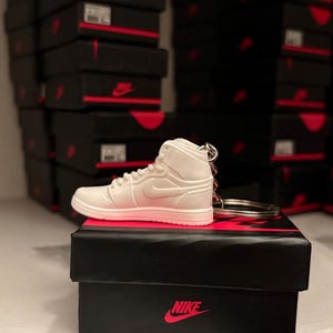 May include: A white high-top sneaker keychain with silver hardware sits atop a black box with a red Nike logo. The background features stacks of black boxes with red accents, suggesting a collection of sneakers.