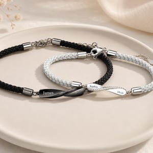 May include: Two matching bracelets with braided bands, one black and one white, are displayed on a plate. Each bracelet has a silver clasp and a decorative center piece engraved with a name. The black bracelet is engraved with the name "James". The white bracelet is engraved with the name "Sophia".
