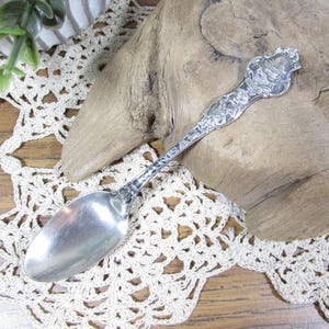 May include: A silver-toned metal souvenir spoon featuring an embossed handle with the text KANSAS. The handle design includes intricate decorative details and a crest at the top, while the bowl of the spoon is smooth, polished, and oval-shaped.