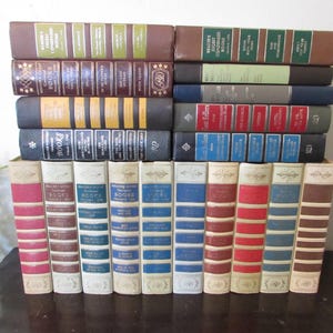May include: A collection of vintage hardcover books with various colors and designs. The spines display titles in gold lettering against backgrounds of red, blue, brown, and white. The books are stacked and arranged, showcasing their decorative covers and aged appearance.