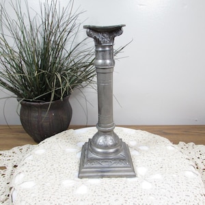 May include: A silver-colored metal candlestick holder with a column design. The candlestick has a square base with ornate detailing and a decorative top. A potted plant and a lace doily are in the background.