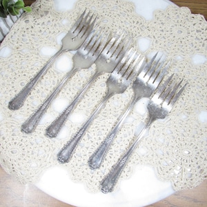 May include: Six vintage silver forks arranged on a cream-colored, crocheted doily. The forks have ornate handles and tines, reflecting light. The doily is placed on a white surface, with a wooden background visible.
