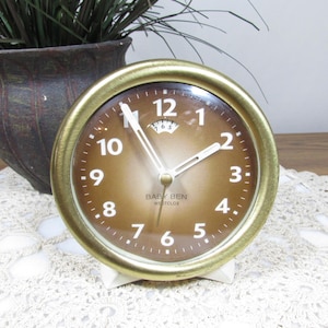 May include: A vintage Baby Ben Westclox alarm clock with a gold-tone metal frame and a brown gradient face. The clock features white numerals and hands, and the text "BABY BEN WESTCLOX" is printed on the face. The clock is set on a white stand.