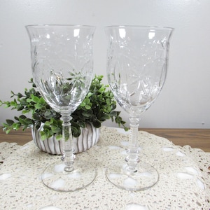 May include: Two clear crystal wine glasses with etched floral designs. The glasses have a classic shape with a stem and a wide base. They are displayed on a white lace doily, with a green plant in the background.
