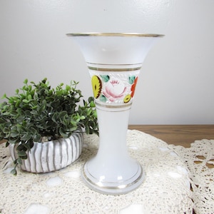 May include: White glass vase with a flared top and a gold rim. The vase features a decorative band with colorful floral designs. A small green plant in a striped pot sits to the left of the vase.