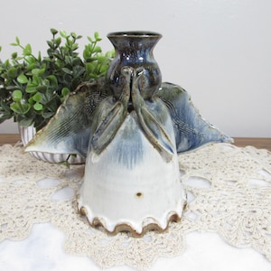May include: Ceramic angel figurine with blue, white, and brown glaze. The angel has outstretched wings and clasped hands. The base is white with a scalloped edge. A small green plant is in the background.