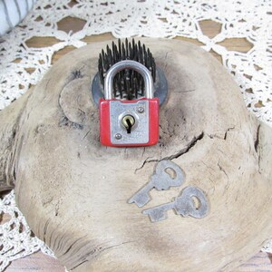 May include: A red and silver padlock with a metallic shackle and a central keyhole, accompanied by two silver-colored metal keys. The padlock is positioned in front of a dark, circular, multi-pronged metal object on a piece of light-colored wood.