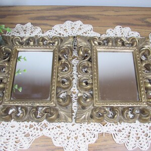 May include: Two rectangular mirrors featuring ornate, metallic gold-toned frames with intricate scrollwork and leaf-like detailing. The mirrors have a smooth, reflective surface and a raised, textured border surrounding the glass panes.