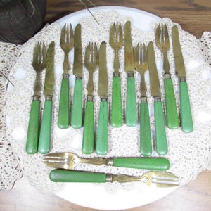 May include: A collection of vintage flatware with green handles and brass-colored blades and tines. The set includes forks, knives, and two specialized utensils. The utensils are arranged on a white plate atop a lace doily.