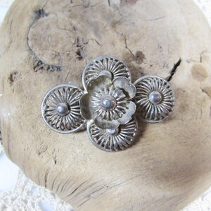 May include: A silver-toned metal brooch in a floral design. The brooch features a central flower with four surrounding circular elements, all with detailed patterns. The brooch is displayed on a piece of weathered wood.