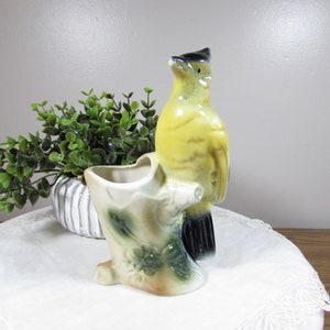 May include: A ceramic planter shaped like a tree stump with a yellow and black bird perched on the side. The stump features molded green leaf details and a hollow opening for plants. The bird has a black head and yellow body with black wings.