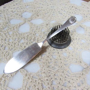 May include: A vintage silver butter knife with a rounded blade and a decorative handle. The handle features a floral design. The knife rests on a black metal flower frog, set against a cream-colored, open-weave doily.