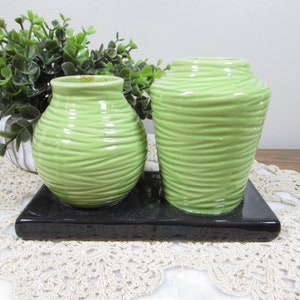 May include: Two light green ceramic vases with a textured, horizontal line design sit on a black rectangular base. The vases are different sizes, one round and one taller. A small green plant in a white pot is in the background.