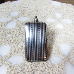 May include: A rectangular silver-toned compact with a vertical striped design and a small, rounded top. The stripes alternate between solid and patterned, creating a textured appearance. The compact is set against a cream-colored, open-weave background.