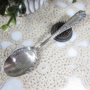 May include: A silver spoon with an ornate handle and a detailed bowl. The spoon is resting on a cream-colored, crocheted doily. A dark, round object with small holes is next to the spoon.