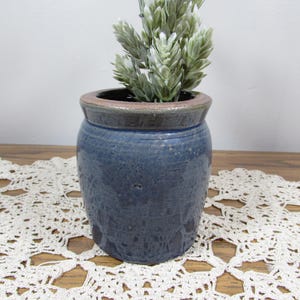 May include: A ceramic crock with a glossy blue glaze and a dark brown rim. The vessel has a rounded body and a wide opening. A small, light green artificial plant with textured, pointed leaves is placed inside the container, extending upwards.