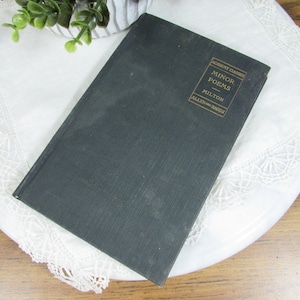 May include: A vintage book titled "Minor Poems" by Milton, with gold lettering on a dark green cover. The book is resting on a white doily with a delicate lace edge. The background includes greenery and a wooden surface.