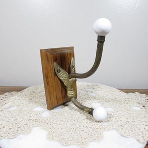 May include: A vintage wall hook with a wooden base and brass eagle detail. The hook has a curved arm with a white ceramic ball at each end. The wooden base is rectangular and has a natural wood grain finish.