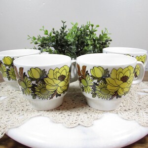 May include: Four white ceramic teacups with yellow floral designs and brown accents. The cups are arranged on a white surface with a lace doily, with a green plant in the background. The cups have a handle.