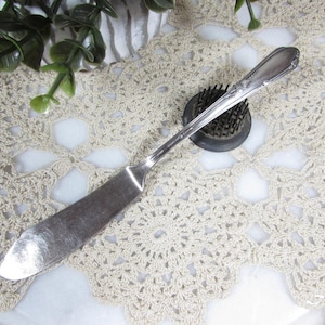 May include: A silver butter knife with a rounded blade and decorative handle rests on a cream-colored, crocheted doily. A dark green floral frog and greenery are also in the image.