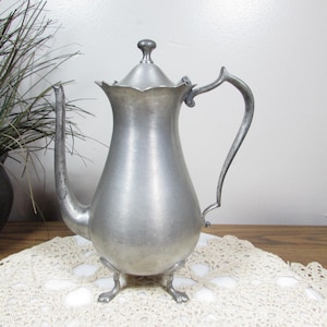 May include: A vintage silver-colored metal teapot with a lid and a curved handle. The teapot has a long spout and sits on three small feet. The teapot is displayed on a white lace doily.