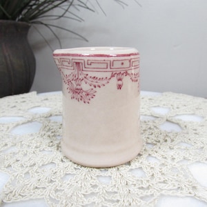 May include: A small, off-white ceramic creamer with a decorative maroon floral and geometric pattern. The creamer has a spout and a cylindrical shape. It is sitting on a white, crocheted doily.