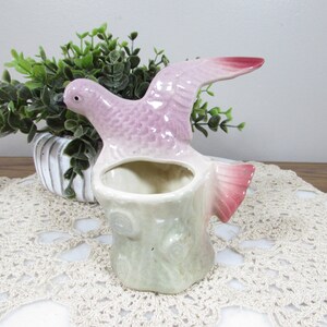 May include: A ceramic bird planter in shades of pink and white. The bird is perched on a tree stump-shaped base, with its wings spread. The planter has a hollowed-out area for holding small plants or flowers. The bird's wings and tail have a darker pink hue.