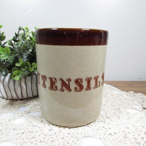 May include: A vintage utensil holder with a brown rim and the word "UTENSILS" in brown lettering. The body of the holder is a light beige color. The holder is cylindrical and sits on a white lace doily.