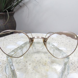 May include: Vintage round eyeglasses with a gold-colored metal frame. The lenses are clear, and the glasses are resting on a clear glass dish. The background includes a decorative lace doily and a dark brown vase with a plant.