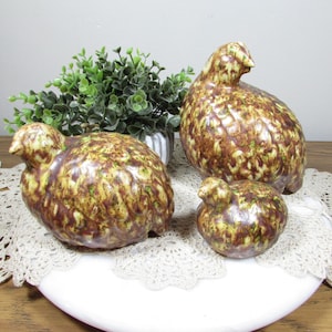 May include: A set of three ceramic bird figurines featuring a mottled glaze in shades of brown, tan, and lime green. The collection includes one large bird, one medium-sized bird, and one small bird, all with smooth, rounded bodies and defined heads.